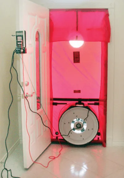 Blower-door test set up in a new home's front doorway: a red fabric panel with a fan and pressure gauge measures air leakage